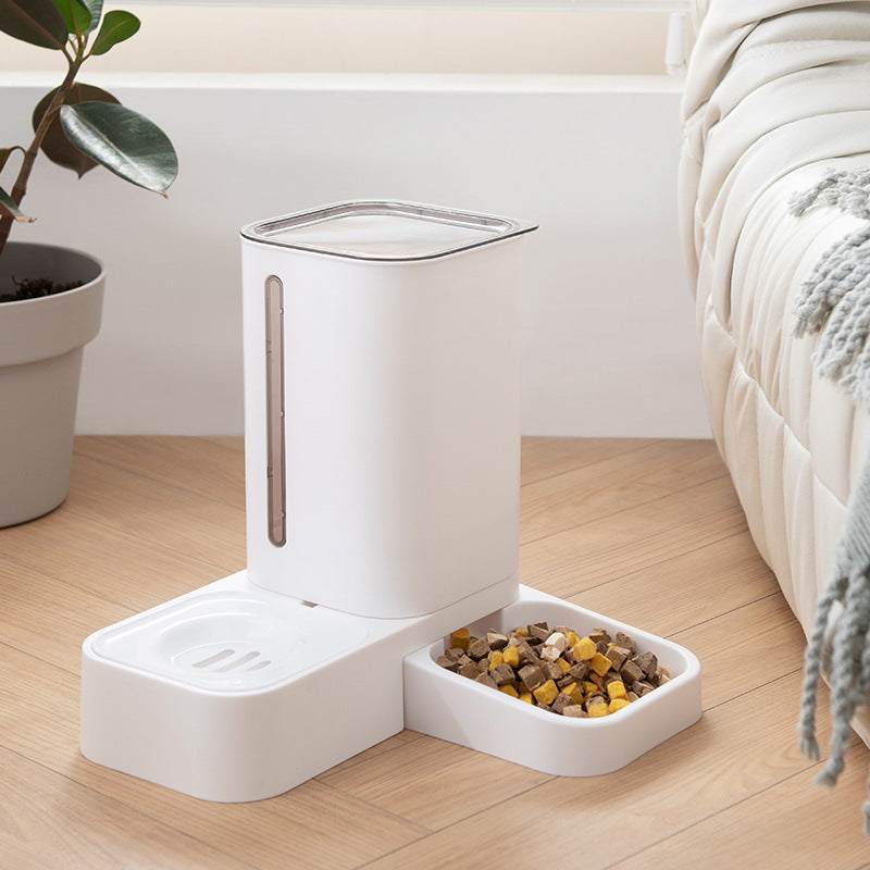 white pet automatic water dispenser with double plastic bowls for food and water on wooden floor next to sofa and houseplant